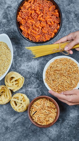 Hand holding a bowl of dry pasta with various types of raw pasta on the marble background. High quality photo